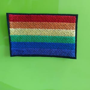 Könnte beinhalten: Ein rechteckiger gestickter Aufnäher mit einem horizontalen Regenbogenflaggen-Design. Der Aufnäher hat einen schwarzen Rand und besteht aus sechs verschiedenen Streifen in Rot, Orange, Gelb, Grün, Blau und Violett, alle mit schimmerndem Garn.