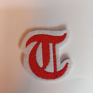 May include: A red embroidered letter "T" patch with a white border. The patch features a classic, bold font style. The patch is likely designed for application on clothing or accessories.