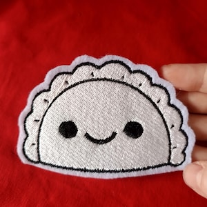May include: A white embroidered patch in the shape of a smiling dumpling with black eyes and a black outline. The patch has a scalloped edge and is set against a red background.