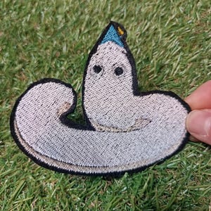 Op de afbeelding: Geborduurde patch met een grillig, lichtgrijs wormachtig wezen met zwarte ogen en een kleine blauwe hoed. De patch heeft een zwarte rand en ligt op groen gras.