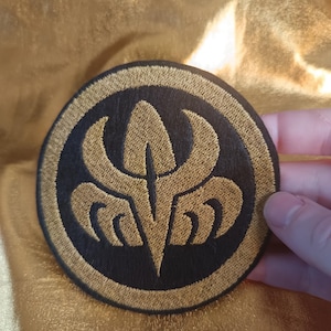 Op de afbeelding: Ronde zwart-gouden geborduurde patch met een gestileerd bloemmotief. De patch heeft een gouden rand en een gouden ontwerp in het midden. De patch wordt door een hand tegen een gouden achtergrond gehouden.