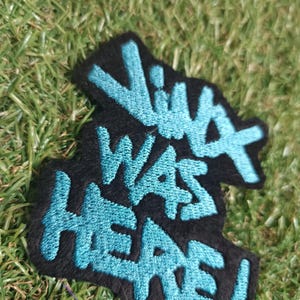 May include: Black patch with the words "VIVA WAS HERE!" embroidered in turquoise. The patch is on a bed of green grass. The text is in a graffiti-style font.