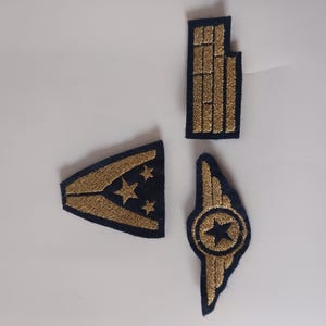 May include: Four navy blue and gold embroidered patches. The designs include a wing with a star, a rectangular shape, and a triangle with stars. These patches could be used for uniforms or crafts.