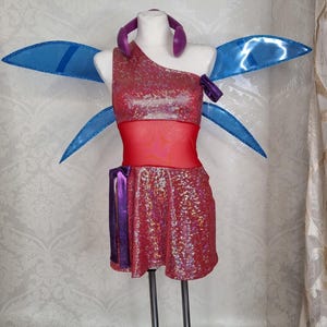 Peut inclure: Un costume rouge et rose à paillettes avec un haut asymétrique, une jupe assortie et un milieu de corps en résille rouge. Le costume comprend des ailes bleues et un serre-tête violet. La tenue est présentée sur un mannequin.