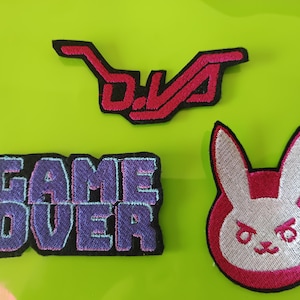 May include: Three embroidered patches featuring gaming themes. One patch displays the pink text D.VA, another shows the purple text GAME OVER, and the third is a white and pink rabbit character icon with a black outline on all three pieces.