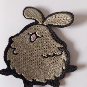 May include: Embroidered patch with a cartoon rabbit design. The rabbit has a fluffy, tan body with black outlines and accents. The ears are tan, and the eye is pink. The patch is suitable for clothing or accessories.