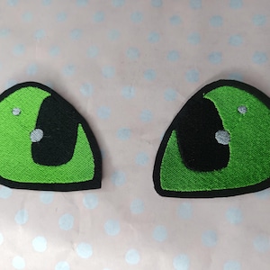 Könnte beinhalten: Zwei aufgestickte Aufnäher in Form von Cartoon-Augen. Jedes Auge hat eine hellgrüne Iris mit einer schwarzen Pupille und einem kleinen weißen Glanzlicht. Die Aufnäher sind schwarz umrandet.
