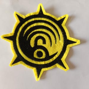 Könnte beinhalten: Gelb-schwarzer, gestickter Aufnäher mit einem Sonnenstrahlen-Design. In der Mitte befindet sich ein stilisiertes schwarzes Symbol mit geschwungenen Linien und einem kreisförmigen Element. Für Kleidung oder Accessoires.