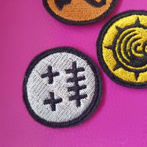 May include: Close-up of three embroidered patches with black borders. One patch is white with black cross-like symbols. Another is yellow with a black sunburst design. The third is orange with a black design.