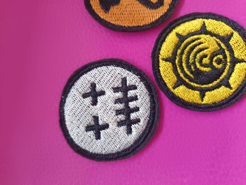 May include: Close-up of three embroidered patches with black borders. One patch is white with black cross-like symbols. Another is yellow with a black sunburst design. The third is orange with a black design.