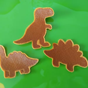 Pode incluir: Três emblemas em forma de dinossauro na cor castanha com uma superfície texturizada e uma borda castanho-clara. O conjunto inclui um dinossauro bípede e dois dinossauros quadrúpedes com placas dorsais, todos com um acabamento têxtil.