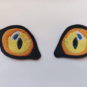 Peut inclure: Deux écussons brodés en forme d'yeux de chat. Chaque œil a un fond noir, un iris orange, un centre jaune, un reflet bleu et une pupille noire. Les écussons sont probablement pour les vêtements ou les accessoires.