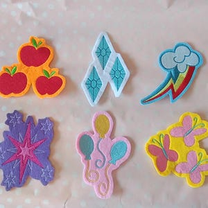 Op de afbeelding: Een set van zes geborduurde viltpatches met kleurrijke ontwerpen: drie rode appels op een oranje achtergrond, drie lichtblauwe ruiten, een regenboogwolk, een paarse ster met kleine sterretjes, drie ballonnen en drie roze vlinders op geel.