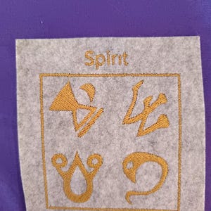 Könnte beinhalten: Ein grauer quadratischer Aufnäher mit goldfarbenen Symbolen und dem Wort "Spirit" in goldfarbenen Buchstaben. Die Symbole sind von einem goldfarbenen Rahmen umgeben. Der Hintergrund ist ein einfarbiger violetter Stoff.