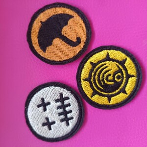 May include: Three embroidered patches with black outlines. One patch features a black umbrella on an orange background. Another has a yellow design on a black background. The third patch has a white background with black cross-like symbols.