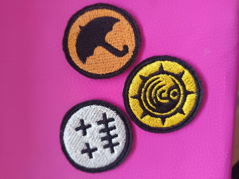 May include: Three embroidered patches with black outlines. One patch features a black umbrella on an orange background. Another has a yellow design on a black background. The third patch has a white background with black cross-like symbols.