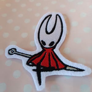 May include: An embroidered patch featuring a stylized character with a white head, black eyes, and a red cape. The character is holding a sword. The patch is white with black and red details, set against a pink polka dot background.