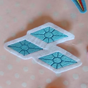 Könnte beinhalten: Weißes Filz-Applikation mit drei türkisfarbenen Diamantformen mit gestickten Details. Die Applikation ist Teil eines größeren Sets an Bastelmaterialien, mit einem Regenbogen und einem rosa-gelben Kreis sichtbar.