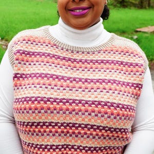 May include: A woman wearing a knitted vest with a geometric pattern in shades of pink, peach, and beige. The vest is layered over a white turtleneck. The background features green grass and trees.