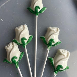 May include: Five white rose-shaped lollipops with green leaves on white sticks. The candies are arranged against a gray background. The lollipops are a sweet treat, perfect for a party or gift.