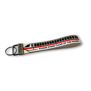 May include: A white fabric keychain with a black and red train design. The keychain has a metal clasp and a ring. The text "PLEASE STAND CLEAR OF THE DOORS" is printed on the keychain.