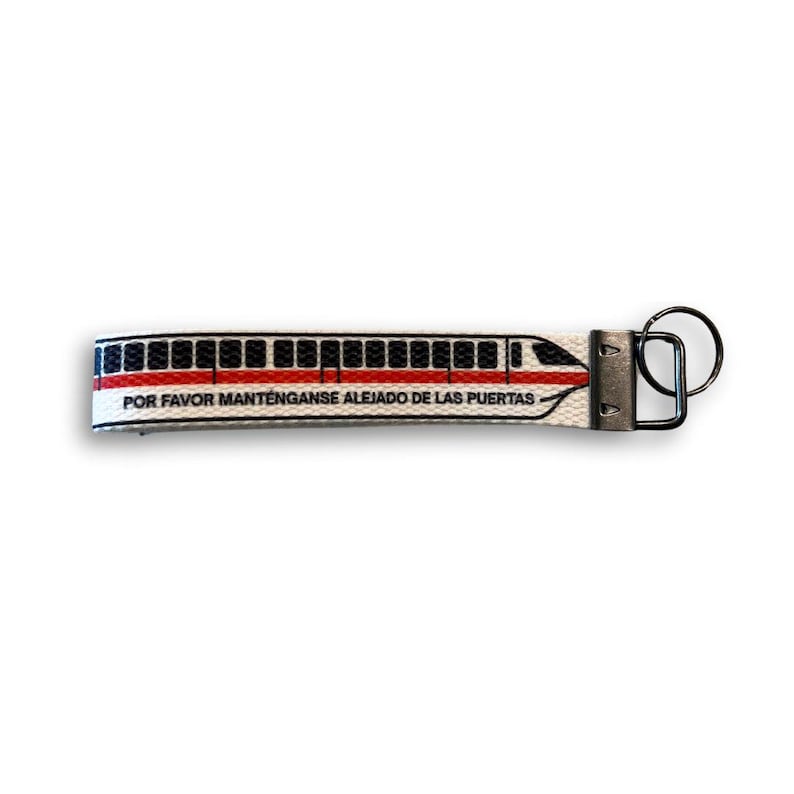 May include: A white keychain with a train design in red and black, and the text "POR FAVOR MANT&Eacute;NGANSE ALEJADO DE LAS PUERTAS". The keychain has a metal clasp and a black keyring.