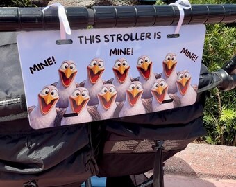 May include: A rectangular white stroller sign featuring a group of cartoon seagulls with orange beaks and wide smiles. The text reads THIS STROLLER IS MINE! and MINE! MINE! The sign is attached to a black stroller handle with two white ribbons.