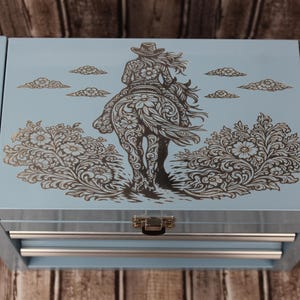 May include: A light blue metal storage box with a top-hinged lid featuring a laser-engraved design of a person riding a horse, surrounded by floral patterns and clouds. The box includes two horizontal drawers with silver-toned metal handles.
