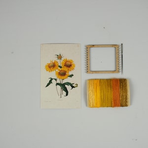 May include: A craft kit for making a small tapestry featuring a vintage illustration of sunflowers. The kit includes a wooden loom, yarn in shades of yellow and orange, and a needle.