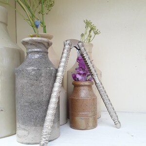May include: Two silver nutcrackers with intricate designs stand against a backdrop of vintage ceramic vases. The vases, in shades of grey and brown, hold flowers. The nutcrackers are positioned upright, slightly angled, adding a touch of elegance.