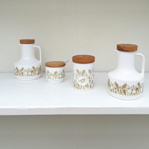 May include: A set of white ceramic kitchen items with wooden lids. The collection includes a jug, a creamer, and two jars. Each piece is decorated with a gold floral design. The items are displayed on a white shelf.