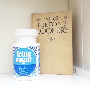May include: A white and blue ceramic container labelled "Icing sugar" with a cake illustration. Next to it is a vintage book titled "Mrs Beeton's Cookery". The items are on a white shelf.