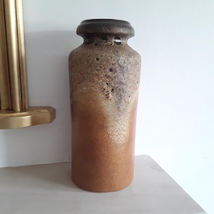 May include: A cylindrical ceramic vase featuring a gradient colour scheme that transitions from a dark brown, textured, and pitted rim to a smooth, lighter tan base. The vase has a distinct, slightly flared neck and a flat, circular opening at the top.