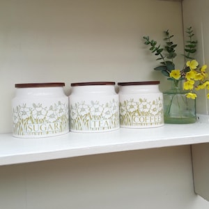 May include: Three white ceramic storage jars with wooden lids, labelled "SUGAR", "TEA", and "COFFEE". Each jar has a floral design in a muted green. The jars are arranged on a white shelf.