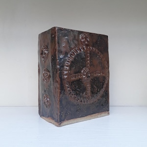 May include: A rectangular, dark brown ceramic vase with a textured surface. The vase features a large, circular gear design in the centre and smaller floral patterns on the sides. The vase is approximately 20 cm tall.