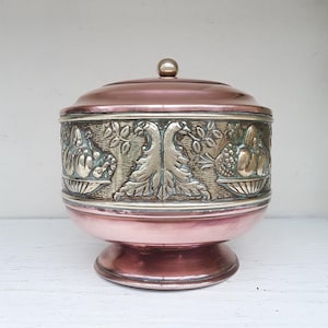 May include: A copper and silver-toned decorative container with a lid. The container has an embossed design of fruits and foliage around the middle section. The lid has a small, round knob. The container sits on a pedestal base.