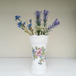 May include: A white porcelain vase with a scalloped rim and embossed detailing at the top. The body features a colourful floral pattern with pink, blue, and green botanical motifs. Several artificial stems with blue and lavender flowers are inside.