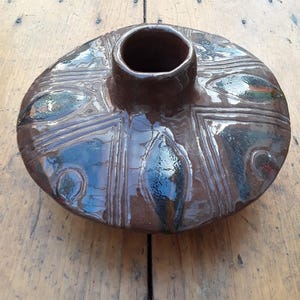May include: A handmade, brown ceramic vase with an oval shape. It has a central, cylindrical opening and is decorated with geometric patterns and dark, glossy accents. The design is rustic and artistic, suitable for home decor.