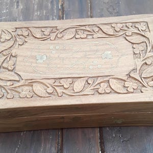 May include: A rectangular wooden box featuring a hand-carved border of leaves and flowers. The centre panel is inlaid with thin brass wire forming a delicate vine pattern with small leaf accents. The natural wood grain is visible throughout the surface.