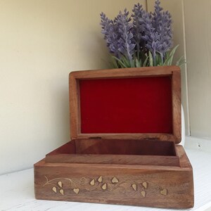 May include: A rectangular wooden box with an open lid, featuring a vibrant red velvet interior lining. The exterior front panel displays an inlaid brass vine pattern with small leaf shapes. The wood grain is visible across the box surface.