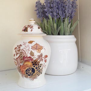 May include: A cream-coloured ceramic ginger jar with a lid, featuring a decorative pattern of autumn-toned leaves, fruits, and a dark blue circular motif. The jar is positioned next to a white ceramic pot containing a bunch of purple lavender flowers.