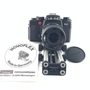 May include: A black Leica R4 camera with a silver lens attached to a black camera tripod. The camera is on a white background. The tripod is made by NOVOFLEX and is made in Germany. The camera has a red dot on the top and the lens has the text "HOFLEXAR 1:4/105 AUTO BELOVE" printed on it.