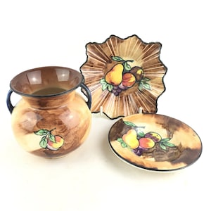 Puede incluir: Un conjunto de tres piezas de cerámica vintage con un diseño de frutas en tonos marrones y crema. El conjunto incluye un jarrón con dos asas, un plato pequeño y un plato más grande con bordes festoneados.