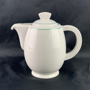 Puede incluir: Cafetera de cerámica blanca con un borde azul claro alrededor de la tapa y el cuerpo. La cafetera tiene un cuerpo redondeado y un asa curva.