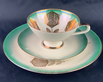 Puede incluir: Un juego de taza y platillo blanco con un diseño floral verde y dorado. La taza tiene un borde dorado y el platillo tiene un borde verde.