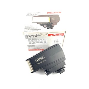 Puede incluir: Un flash de cámara negro Metz mecablitz 28 AF-4 M con un reflector plateado y dorado. El flash está inclinado hacia el espectador, con el logotipo de Metz visible. La caja y el embalaje del producto también son visibles.