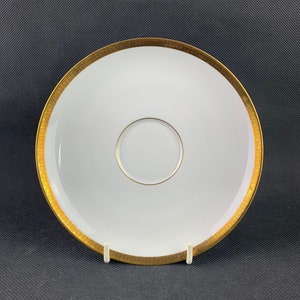 Könnte beinhalten: Eine weiße Untertasse mit goldfarbenem Rand und einem goldfarbenen Mittelkreis. Die Untertasse ist aus Porzellan und hat eine glatte, glänzende Oberfläche.