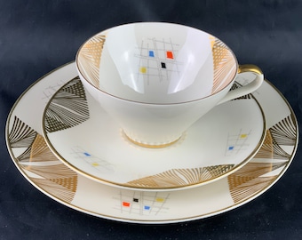 Puede incluir: Un juego de taza y platillo de té blanco con un diseño geométrico dorado. La taza y el platillo tienen un patrón de líneas dorées y cuadrados con acentos rojos, azules y amarillos.
