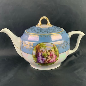 Könnte beinhalten: Eine blaue und weiße Porzellan-Teekanne mit goldener Verzierung und einem gemalten Bild von zwei Frauen und einem Kind in einem Garten. Die Teekanne hat einen runden Körper und einen Deckel mit einem Knopf.
