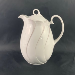 Può includere: Teiera in porcellana bianca con un delicato motivo floreale rosa e un manico curvo.